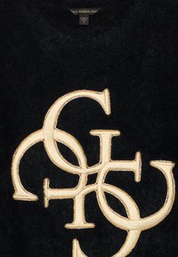 Εμπλεγμένα κρεμ χρώματος "G" και "S" κεντημένα σε μαύρο μαλακό ύφασμα με μικρές χρυσές λεπτομέρειες από χάντρες, ετικέτα μάρκας GUESS από πάνω.