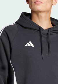 Primo piano di una persona che indossa un hoodie nero Adidas con logo bianco, cordini e dettagli a strisce bianche sulla spalla e sul lato.