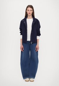 Navy zip-up jacket with a high collar, worn over a white long-sleeve shirt, paired with wide-legged blue denim jeans and cream shoes.