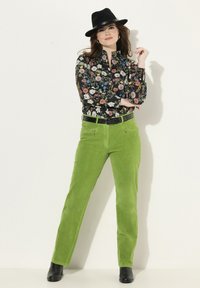 Blus med blommönster på svart bakgrund, ihop med livfulla gröna kordbyxor och en svart fedora-hatt. Svart bälte avslutar looken.