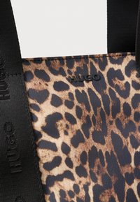 Torba tote w leoparda, wykonana z gładkiego materiału, z czarnymi detalami logo i trwałymi czarnymi paskami, podkreślająca odważny zwierzęcy wzór.