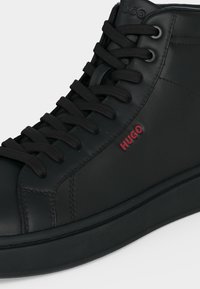 Baskets montantes en cuir noir avec lacets noirs et logo rouge "HUGO" sur le côté, avec une semelle épaisse en caoutchouc noir.