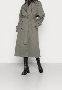 Szary długi trenchcoat z podwójnym zapięciem, wiązanym w pasie oraz bocznymi kieszeniami, w połączeniu z czarnymi botkami na obcasie i legginsami. Gładka faktura materiału.