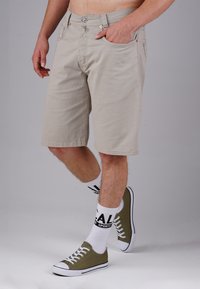 Beige shorts with a straight cut, five pockets, and metal button closure. Worn with white sport socks and olive green sneakers.