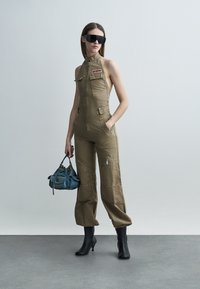 Szczupła kobieta w oliwkowozielonym kombinezonie na zamek, czarne botki za kostkę, duże czarne okulary przeciwsłoneczne, trzymająca niebieską torebkę, stojąca na szarym podłożu.