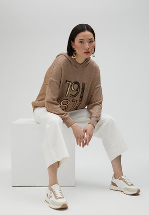 Haut à capuche en bronze pailleté avec du texte noir et doré, un pantalon blanc court, et des baskets blanches avec des accents beige, assis sur un piédestal blanc.