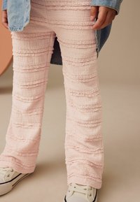Helle rosa ausgestellte Hosen mit strukturierten vertikalen Streifen und Spitzenakzenten, kombiniert mit weißen Turnschuhen mit schwarzer Umrandung und Schnürsenkeln.