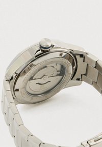 Stainless steel watch back showing automatic movement, visible gears, a crown, and linked metal bracelet against a light background.