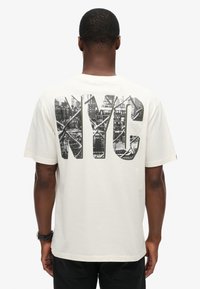Camiseta de algodón crema con un gran gráfico negro de NY en la parte trasera, que presenta un detalle del horizonte de la ciudad. Mangas cortas y un ajuste relajado.