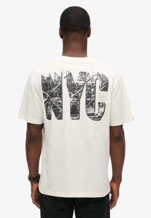 Camiseta de algodón crema con un gran gráfico negro de NY en la parte trasera, que presenta un detalle del horizonte de la ciudad. Mangas cortas y un ajuste relajado.