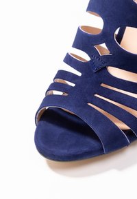 Marinblå mocka klacksandaler med öppen tå, utrustade med flera utskurna remmar och en slät sula som betonar textur och färgkontrast.