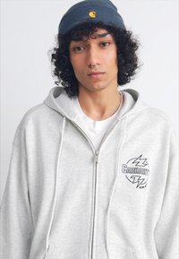 Lys grå zip-opp hettegenser med lommer foran og svart Carhartt-logo. Brukt over en hvit skjorte, kombinert med en mørk lue. Myk stoffoverflate.