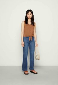 Top castanho, sem mangas, com botões à frente e laço; jeans azuis, boca de sino; chinelos pretos; saco pequeno de cor clara. Fundo simples.