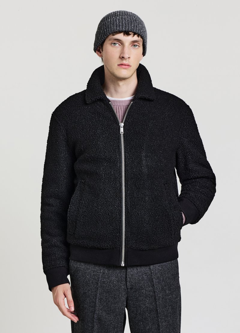 Schwarze Lammfelljacke mit Reißverschluss, Kragen und gerippten Bündchen, getragen über einem hellrosa Pullover. Das Modell trägt außerdem eine graue gestrickte Mütze.