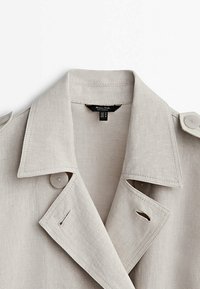 Jasnoszary płaszcz trench z lnu, z wyciętym kołnierzem, podwójnymi guzikami i bocznymi kieszeniami. Zauważalne detale przeszyć wzdłuż szwów.