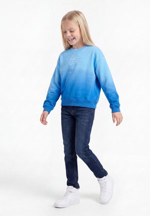 Lächelndes Mädchen läuft, trägt ein blaues Sweatshirt mit Farbverlauf, dunkle Jeans und weiße High-Top-Sneaker vor weißem Hintergrund.