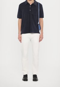 Polo de manga corta en color azul marino con cuello y bolsillo, combinado con jeans blancos y zapatos negros. El modelo sostiene un pequeño bolso azul.