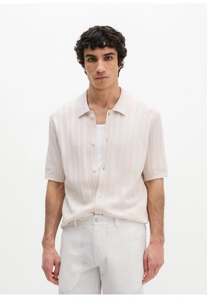 Junger Mann mit lockigem dunklem Haar, der ein hellbeiges kurzärmeliges Hemd über einem weißen Tanktop und weißen Hosen trägt und vor weißem Hintergrund steht.