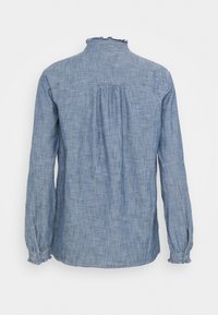 Chemise en denim bleu clair avec des manches longues, un col volanté et un pli au dos. Le tissu a une apparence douce et légèrement texturée.