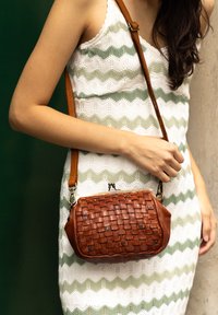 Sac bandoulière en cuir tressé marron avec une forme structurée, dotée d'un fermoir en argent et d'une bandoulière beige. Porté avec une robe à motifs.