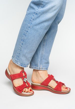 Feet wearing red open-toe sandals with two adjustable straps and decorative metal accents, paired with light blue denim jeans.