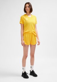 Gult sportset med tröja och shorts, tillverkat i lätt tyg. Har rund halsringning, logotyp på tröjan och randiga detaljer på shortsen.