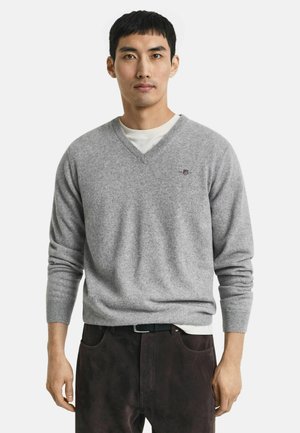 Grauer Woll-Pullover mit V-Ausschnitt, gerippten Bündchen und Saum, mit einem kleinen bestickten Logo auf der Brust. Wird über einem weißen T-Shirt getragen.