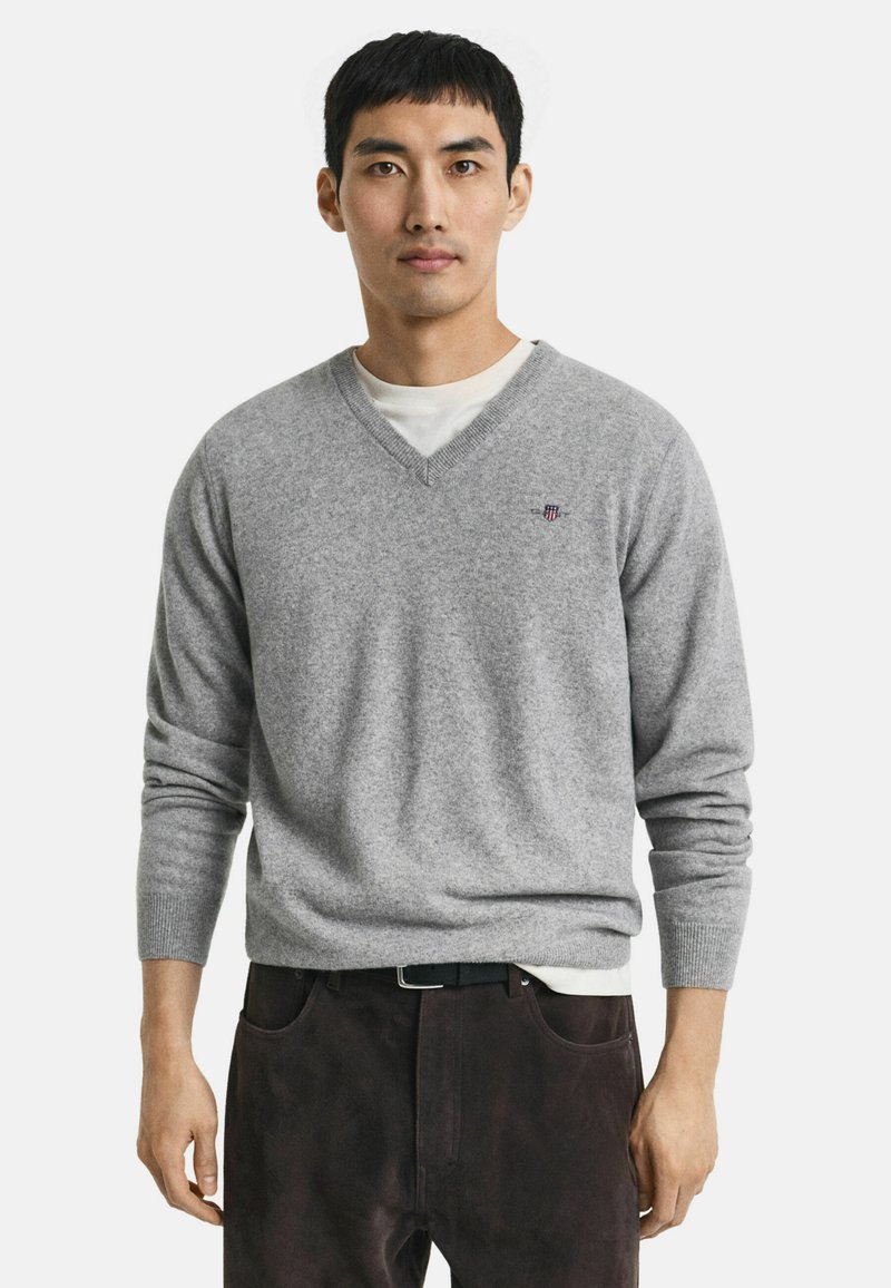 Maglione grigio in lana a scollo a V con polsini e orlo a coste, con un piccolo logo ricamato sul petto. Indossato sopra una maglietta bianca.