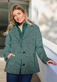Femme souriante avec de longs cheveux blonds, portant une veste à boutons texturée verte et un gilet matelassé vert, appuyée sur un rebord blanc en extérieur.