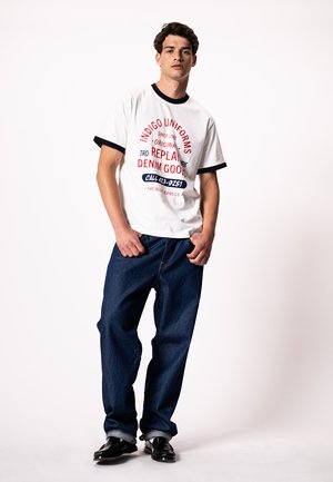Mladi moški stoji oblečen v belo majico s črnim robom in grafičnim potiskom, široke temno modre kavbojke ter črne čevlje na belem ozadju.