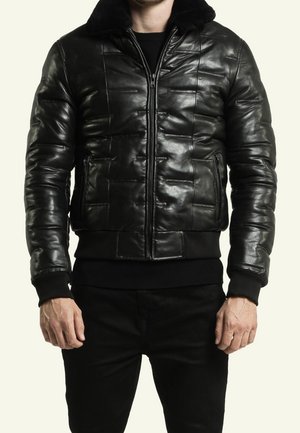Mann trägt eine schwarze gesteppte Lederjacke mit Frontreißverschluss, seitlichen Reißverschlusstaschen und einem schwarzen Pelzkragen, kombiniert mit schwarzen Hosen.