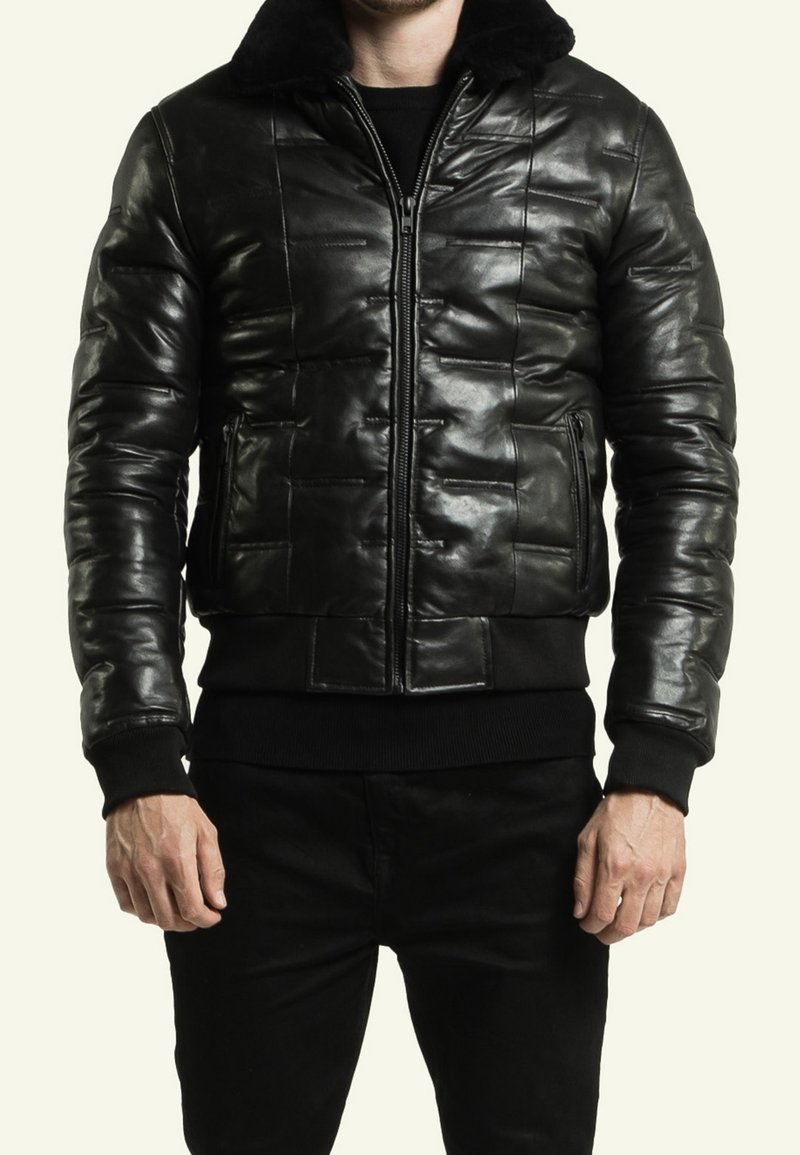 Homme portant une veste en cuir matelassée noire avec une fermeture éclair frontale, des poches latérales zippées et un col en fourrure noire, associée à un pantalon noir.