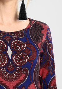 Hedvábný top s vzorem v námořnické modré, červené a bílé barvě, zdobený složitými paisley grafikami. Hladká tkanina a kulatý výstřih zdůrazněný detaily s třásněmi.