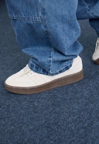 Person wearing blue jeans and white sneakers with brown soles standing on dark blue carpeted floor.