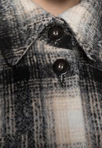 Plaid flannel skjorte i sort, grå og creme med en struktureret overflade. Har fremtrædende sorte knapper ved kraven og brystet.