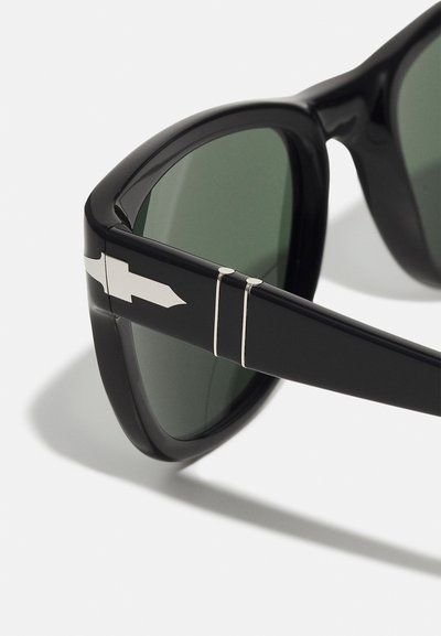Óculos de sol pretos com lentes verdes. Armação texturizada e arredondada com acentos em forma de seta prateada nas hastes.