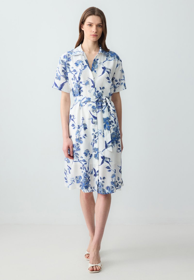 Robe à manches courtes en tissu blanc avec des motifs floraux et d'oiseaux bleus. Dispose d'un col et d'un détail de taille nouée.