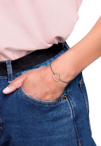 Goldfarbener Armreif mit einem strukturierten runden Anhänger an einem dünnen Drahtband, getragen an einer Hand, kombiniert mit einem rosa Oberteil und einer blauen Jeans.