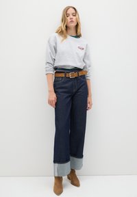 Giovane donna in piedi contro un muro bianco, indossa una felpa grigia, jeans larghi scuri con risvolti, cintura marrone e stivaletti in suede color beige.