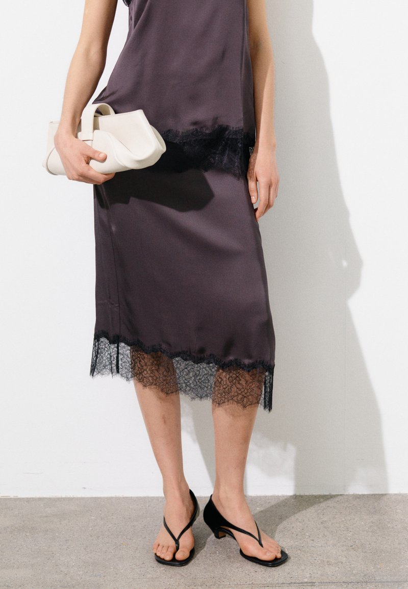 Person trägt ein violettes Seidenkleid mit schwarzem Spitzenabschluss, hält eine weiße Clutch und trägt schwarze, riemchenbesetzte Sandaletten mit Absatz.