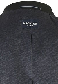 Zwarte jas met een subtiel patroon, voorzien van een gestikt label met de tekst "HECHTER PARIS." Gladde textuur en een gestructureerde kraag.
