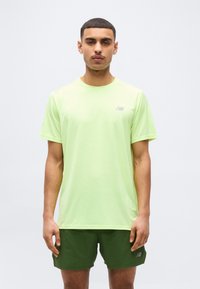 Άνδρας που στέκεται, φορόντας ένα ανοιχτοπράσινο t-shirt New Balance και σκούρα πράσινα σορτς New Balance, με φόντο ένα απλό ανοιχτό χρώμα.