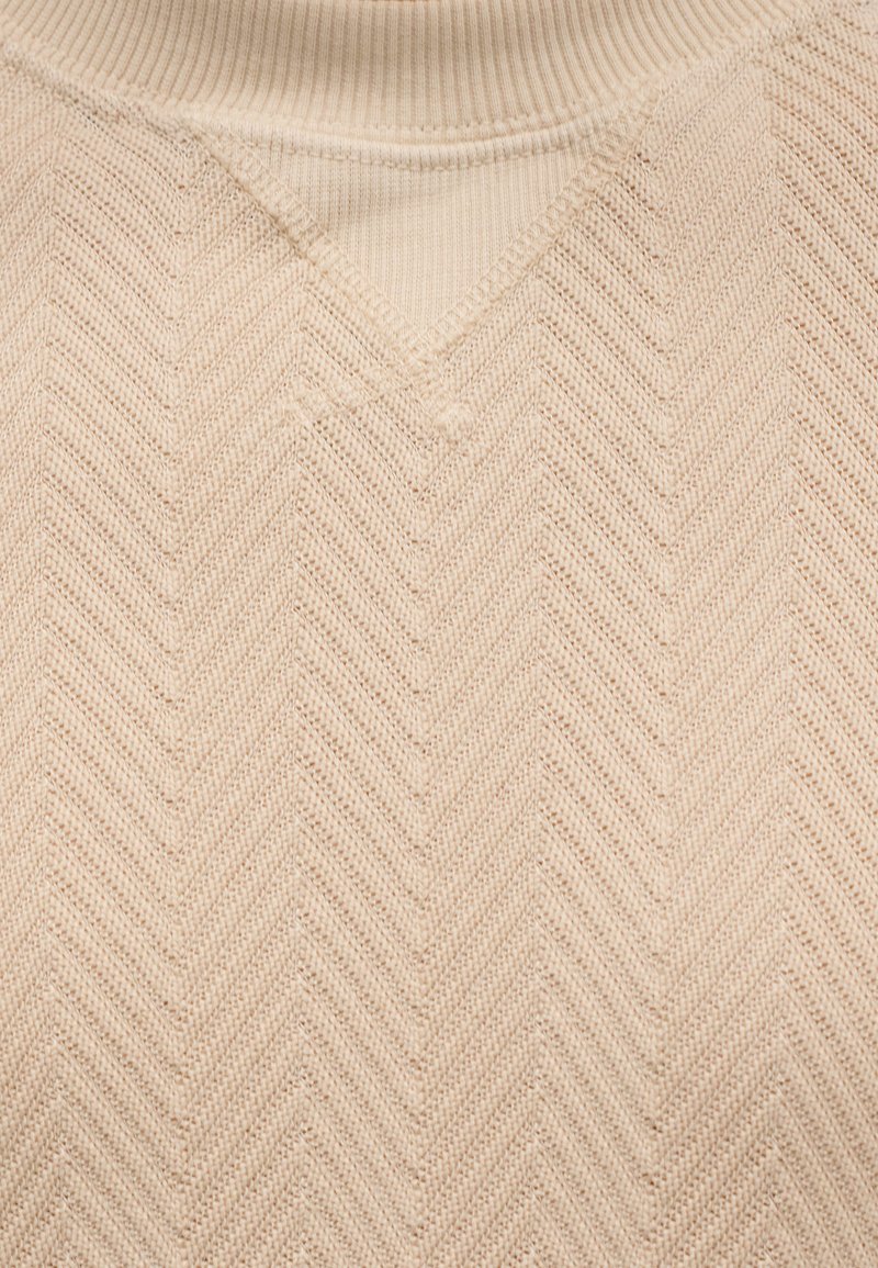 Pull en maille beige clair avec un col en V et un motif en chevrons. La texture est douce et côtelée autour du col.