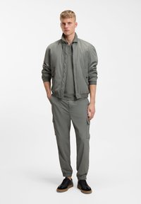 Jeune homme portant une veste bomber grise, un pantalon cargo assorti et des baskets noires, debout les mains dans les poches sur un fond blanc.