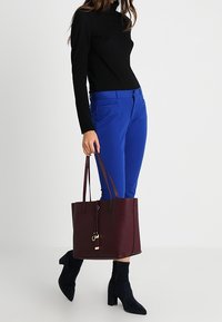 Borsa a tote bordeaux realizzata in materiale sintetico con una texture liscia, dotata di manici lunghi e un dettaglio di portachiavi appeso. Pantaloni blu brillante e top nero.