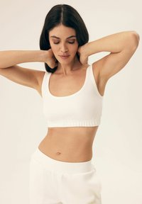 White crop top made of soft fabric with a rounded neckline and wide straps, featuring a gathered hem for a snug fit. Paired with white bottoms.