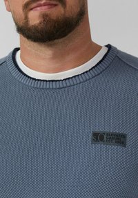 Brodaty mężczyzna noszący niebieski sweter o fakturze z białą koszulką pod spodem, z czarną łatą, na której widnieje napis "S.O Elevated Casual Est. 1969."