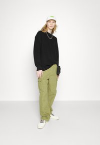 Schwarzer Sweatshirt, grüne Cargo-Hose mit Seitentaschen, weiße Schuhe und eine weiße Mütze mit grünem Logo, stehend vor einem hellen Hintergrund.