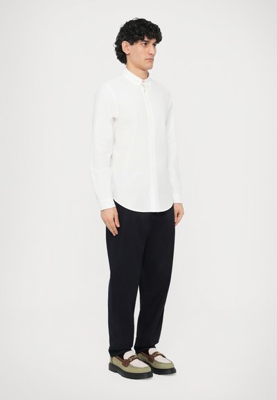 Camicia bianca a maniche lunghe con bottoni, pantaloni larghi scuri e scarpe bicolore con tonalità verde e marrone, dotate di suola spessa.