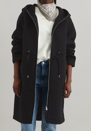 Manteau noir à capuche avec fermeture éclair à l'avant, taille ajustable par cordon, et poches latérales. Présente des manches longues et une coupe décontractée. Tissu texturé.